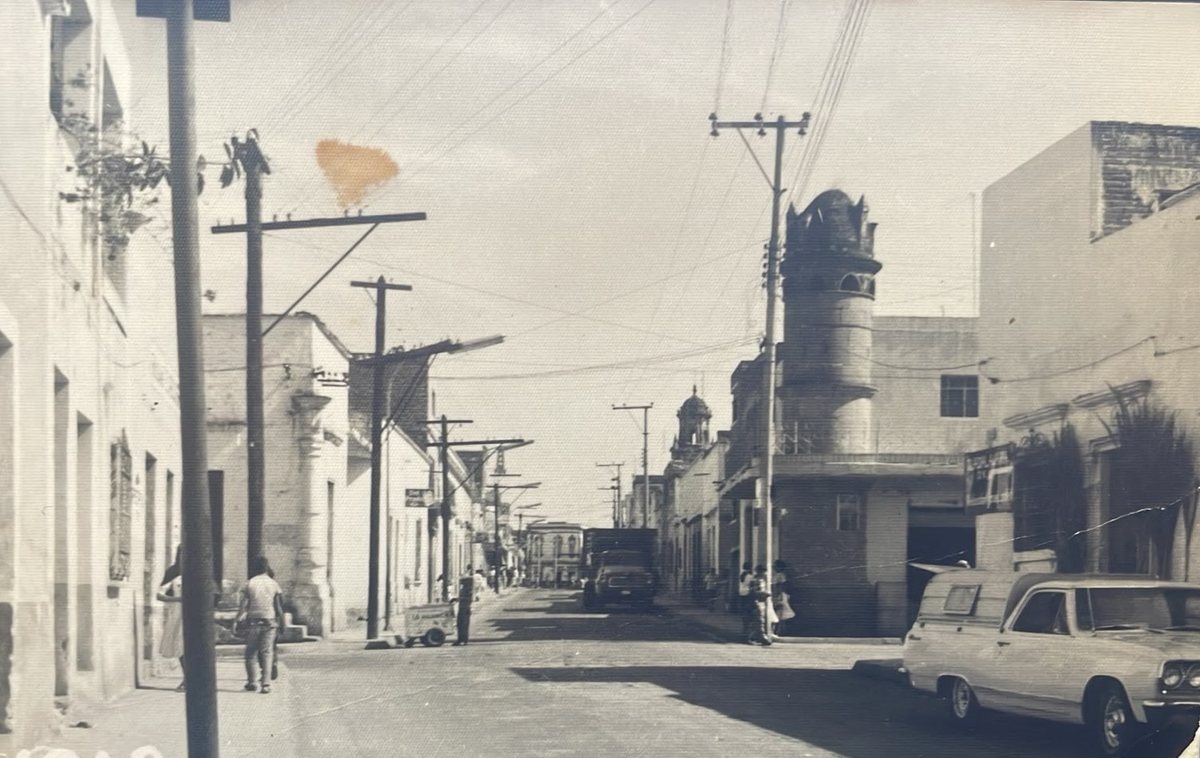 Fotografía antigua en sepia de una calle de Zapotlanejo: casas bajas, postes de luz, una torre al fondo y un coche de la época estacionado.