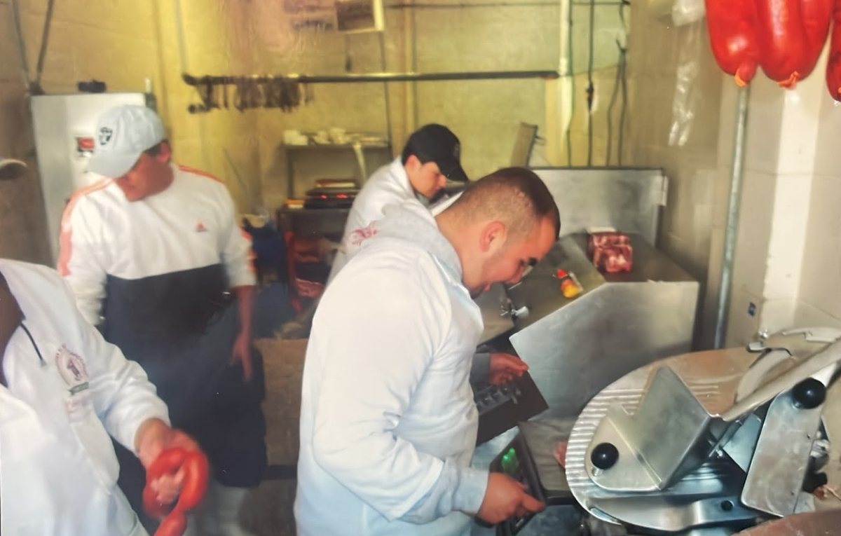 Fotografía antigua a color: el equipo, de blanco, trabajando en la carnicería junto a la rebanadora, con chorizos colgados al fondo.