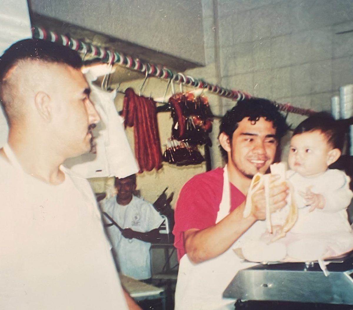 Fotografía antigua a color: en el mostrador, un carnicero sonriente sostiene a un bebé mientras otro hombre los mira; detrás cuelgan chorizos y una guirnalda tricolor.
